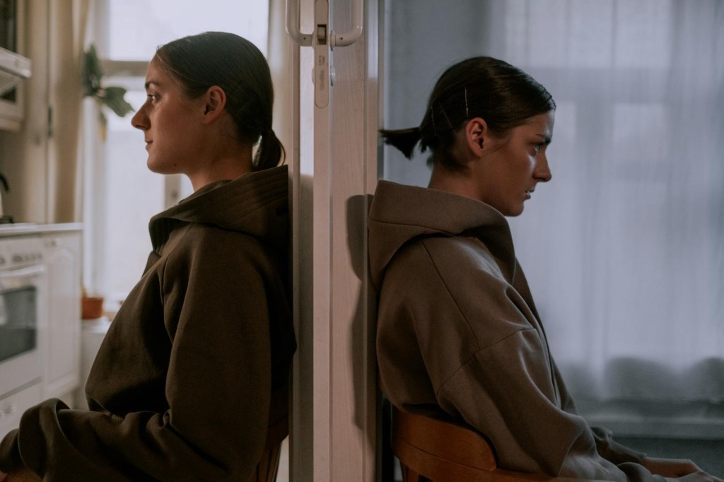 Image of a woman sitting next to a mirror showcasing two versions of herself and her multilayer personality. One face is smiling and the other is deep in thought. 